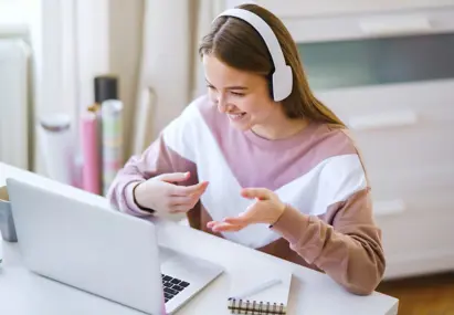 Vrouw met koptelefoon voert online gesprek via laptop aan bureau, glimlachend en gebarend tijdens videogesprek.
