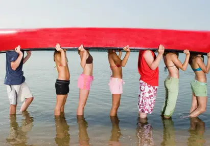 Groep mannen en vrouwen in badkleding houden een omgekeerde kano vast die hun hoofden verbergt