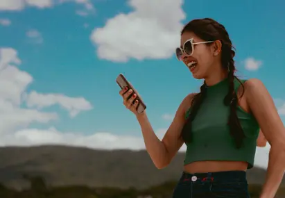Vrouw met twee vlechten, zonnebril en groen shirt kijkt lachend naar haar telefoon
