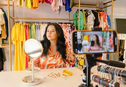 Vrouw presenteert sieraden en filmt zichzelf met smartphone in kleurrijke kledingwinkel met rekken vol felgekleurde kleding.