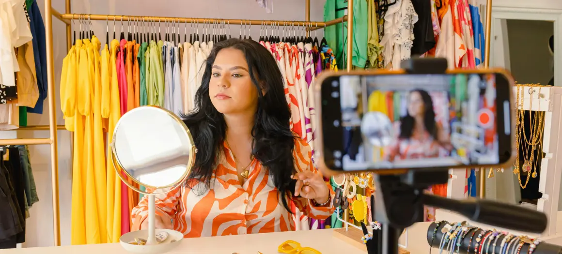 Vrouw presenteert sieraden en filmt zichzelf met smartphone in kleurrijke kledingwinkel met rekken vol felgekleurde kleding.